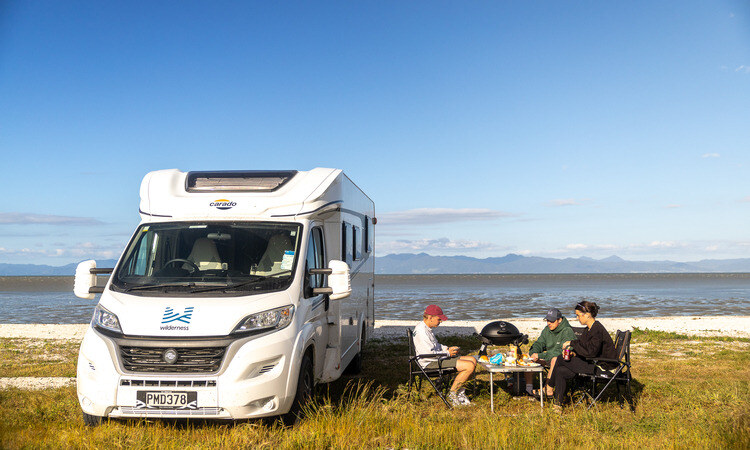 A group of friends sitting with camping table and chairs provided by Wilderness Motorhome while enjoying barbecue outside of their motorhome